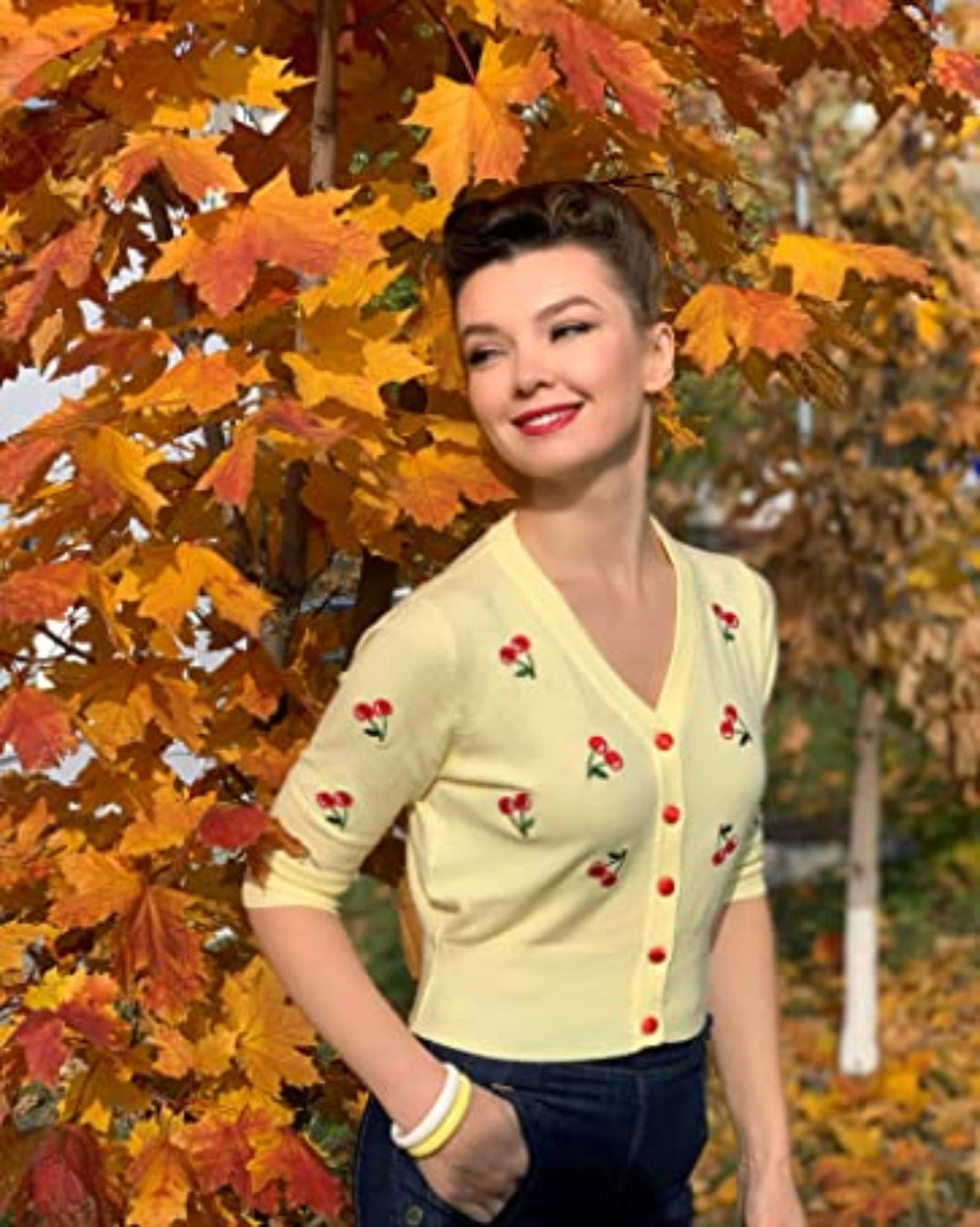 Woman in a cropped cherry cardigan posing among autumn leaves, showcasing a vintage style and playful charm.