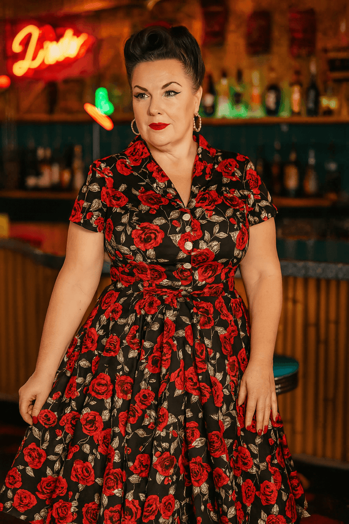 Woman wearing Rose Romance Vintage Swing Dress with red floral print, showcasing a retro style in a vibrant bar setting.