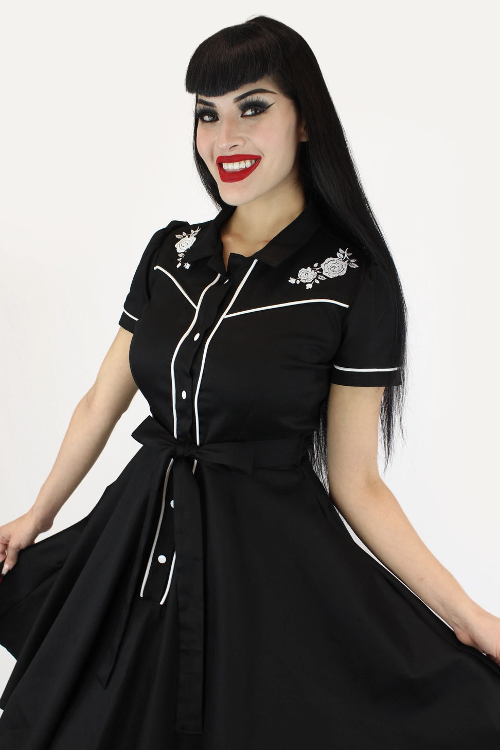 A woman modeling a black short-sleeve swing dress with white piping, floral embroidery on the chest, and a matching waist tie, set against a plain white background.