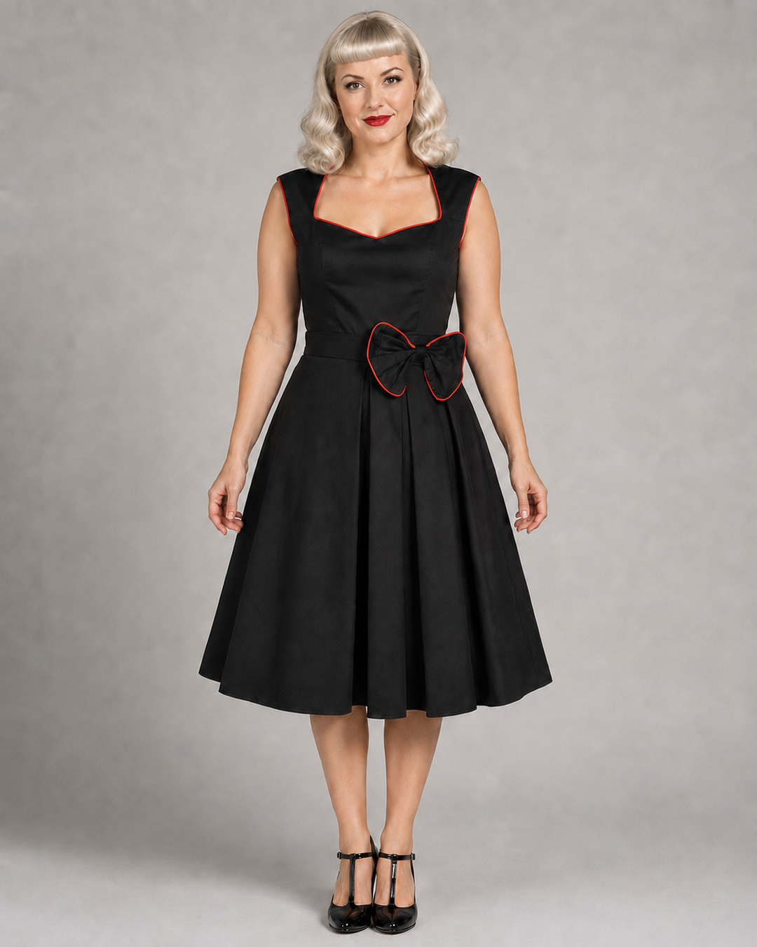 A woman modeling a black sleeveless swing dress with a sweetheart neckline, a matching black belt, and a large black bow with red trim at the waist, photographed against a neutral gray studio background.