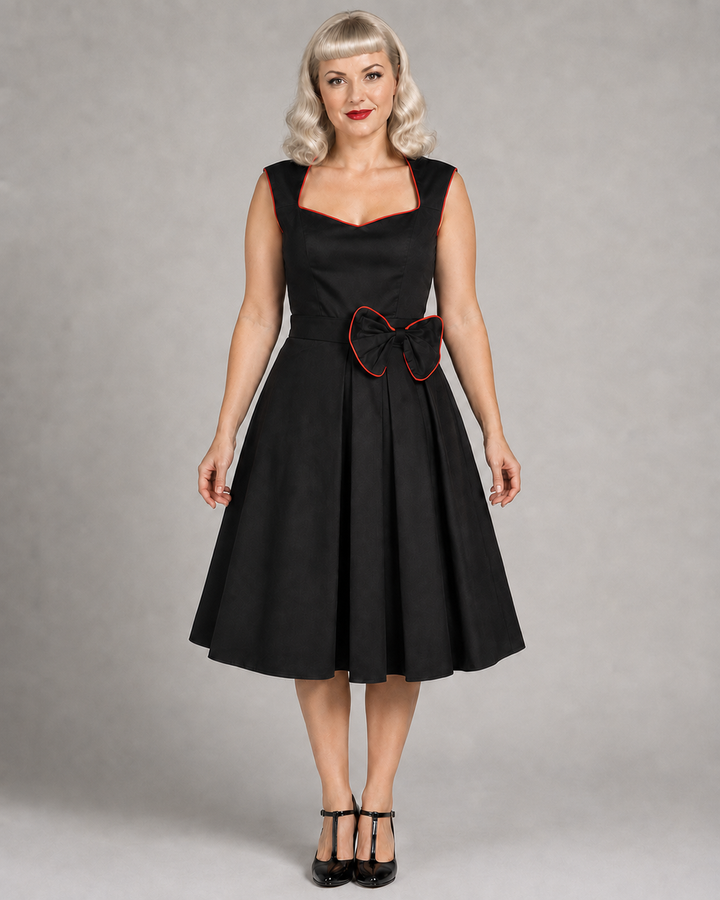 A woman modeling a black sleeveless swing dress with a sweetheart neckline, a matching black belt, and a large black bow with red trim at the waist, photographed against a neutral gray studio background.