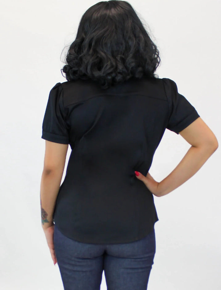 A woman viewed from behind wearing a black short-sleeve top with shoulder cutouts and dark blue jeans, standing against a plain white background.