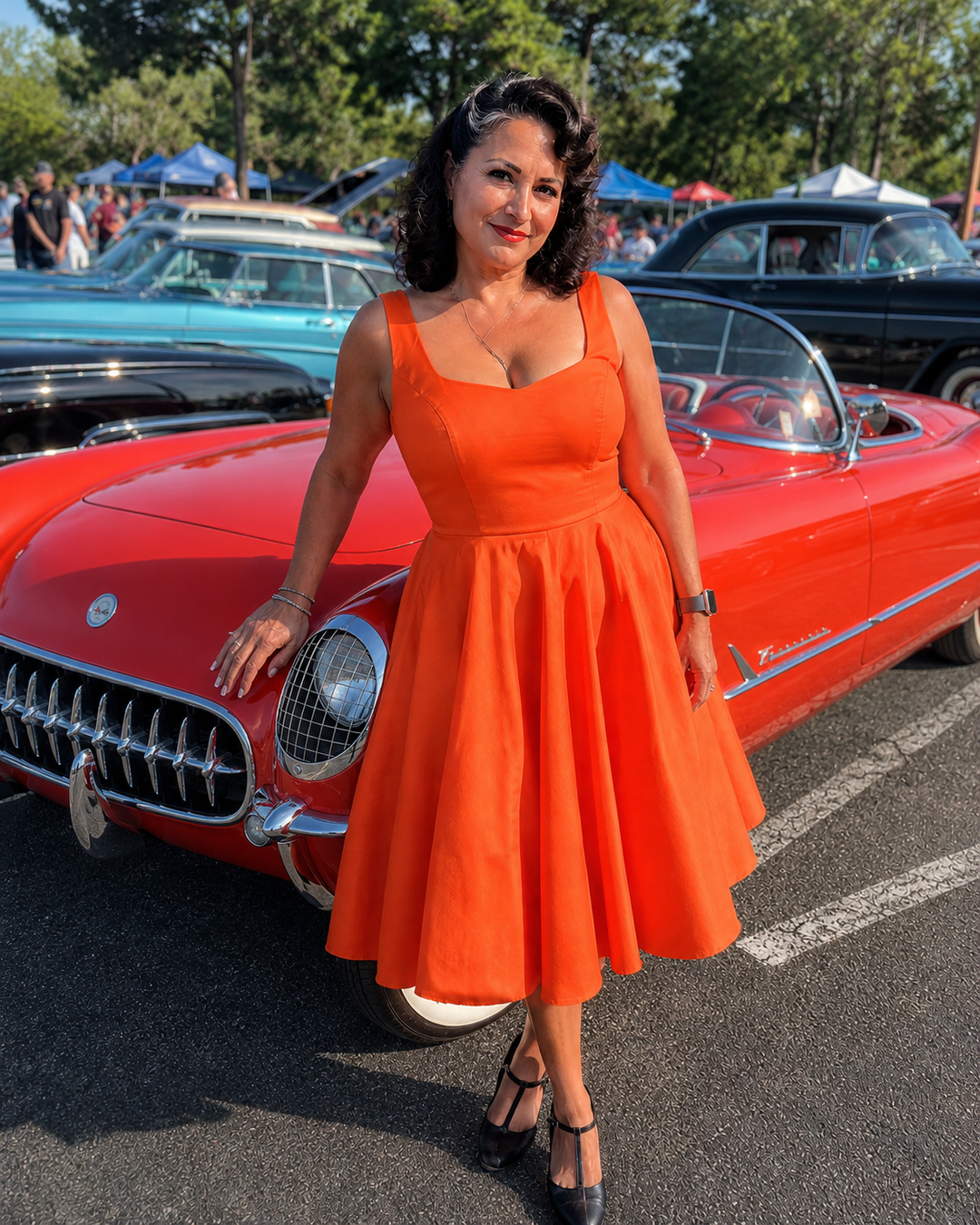 A woman wearing a bright orange sleeveless swing dress stands in an outdoor car show parking lot next to a red classic car.