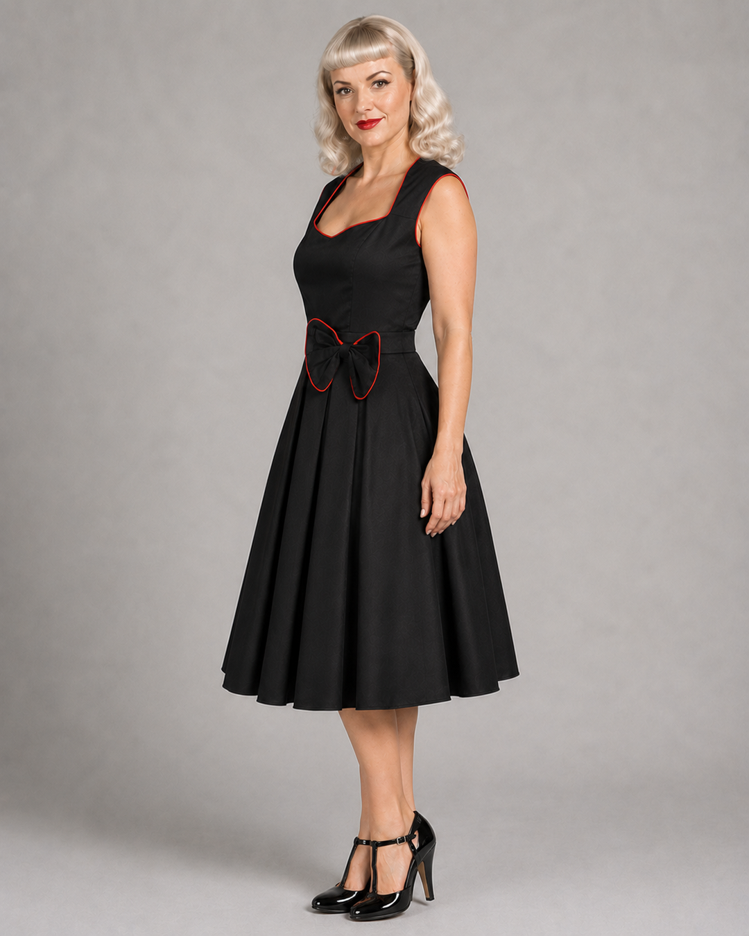 A woman modeling a black sleeveless fit-and-flare dress with a square neckline, full skirt, and a red bow detail at the waist, photographed against a plain gray studio background.