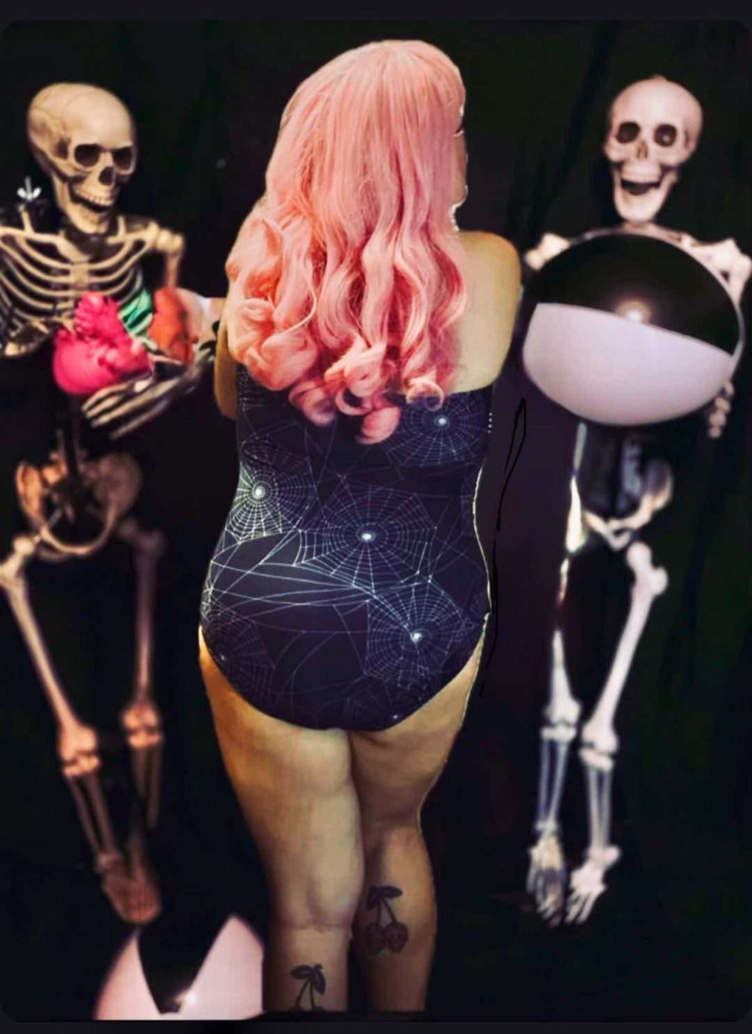 Person with pink hair wearing a dark blue geometric-patterned swimsuit in front of skeletons and a black sphere.