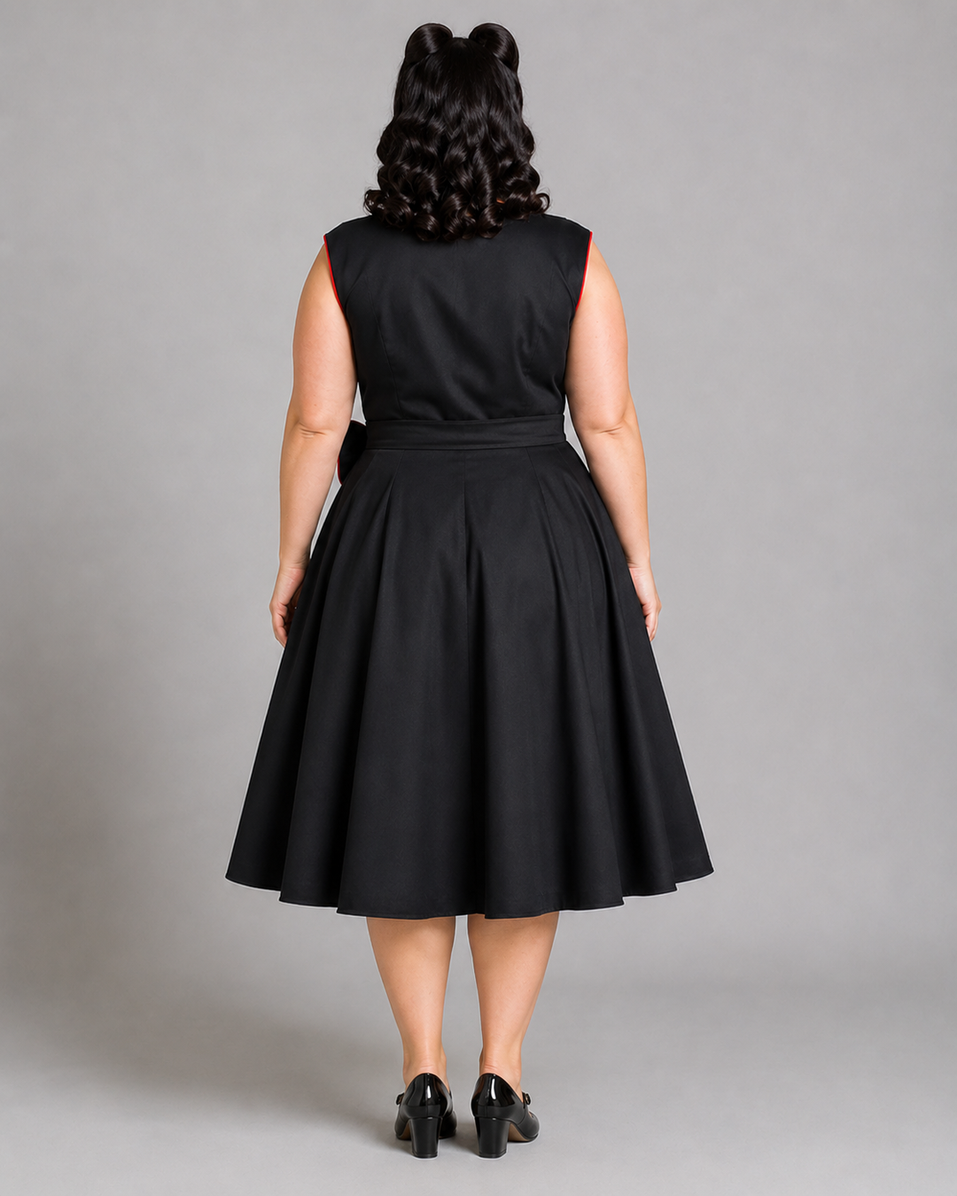 A woman viewed from the back wearing a sleeveless black A-line dress with a defined waist and flared skirt, paired with black heels, against a plain gray studio background.