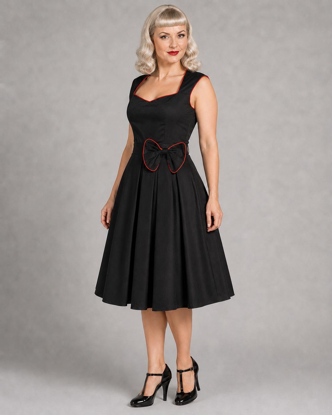 A woman modeling a black sleeveless swing dress with a sweetheart neckline, pleated skirt, and a bow detail at the waist outlined in red trim, photographed against a neutral gray studio background.