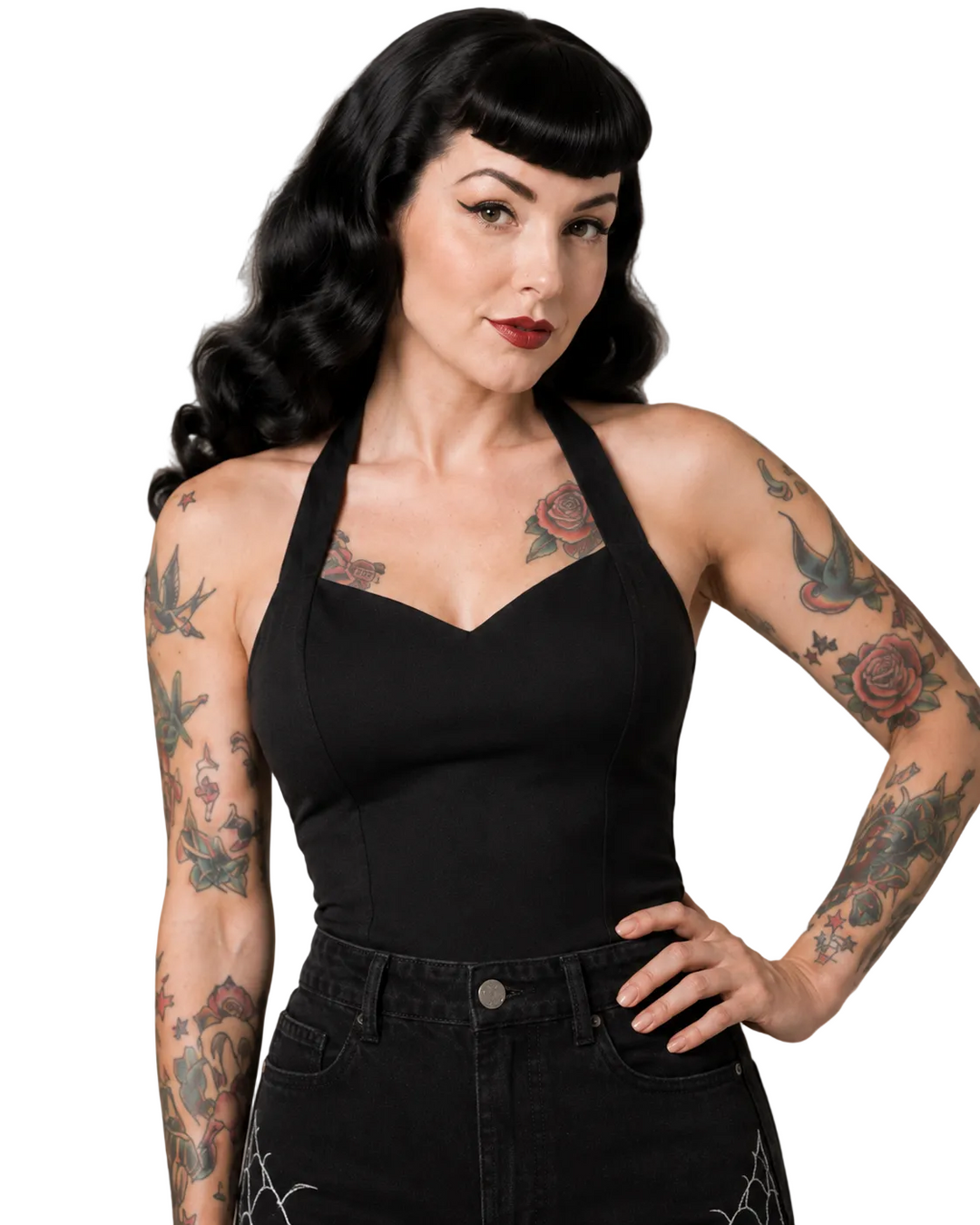 Woman wearing a black halter top and black pants with tattoos on her arms against a neutral background