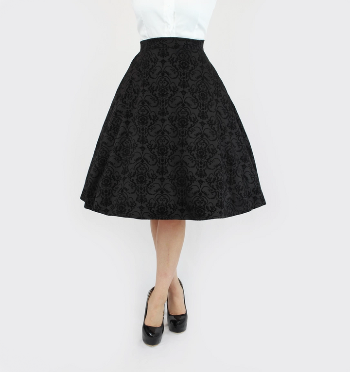 A black high-waisted A-line skirt with a textured damask brocade pattern, shown on a model wearing a white top and black heels against a plain white background.