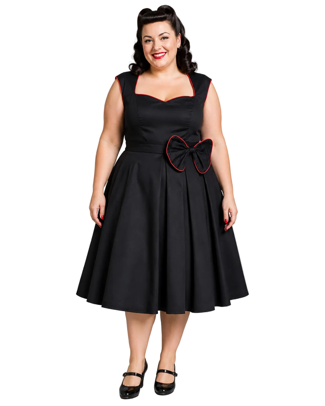 A woman modeling a black 1950s-style swing dress with red piping trim, a sweetheart neckline, and a large bow detail at the waist, paired with black Mary Jane shoes against a solid gray studio background.