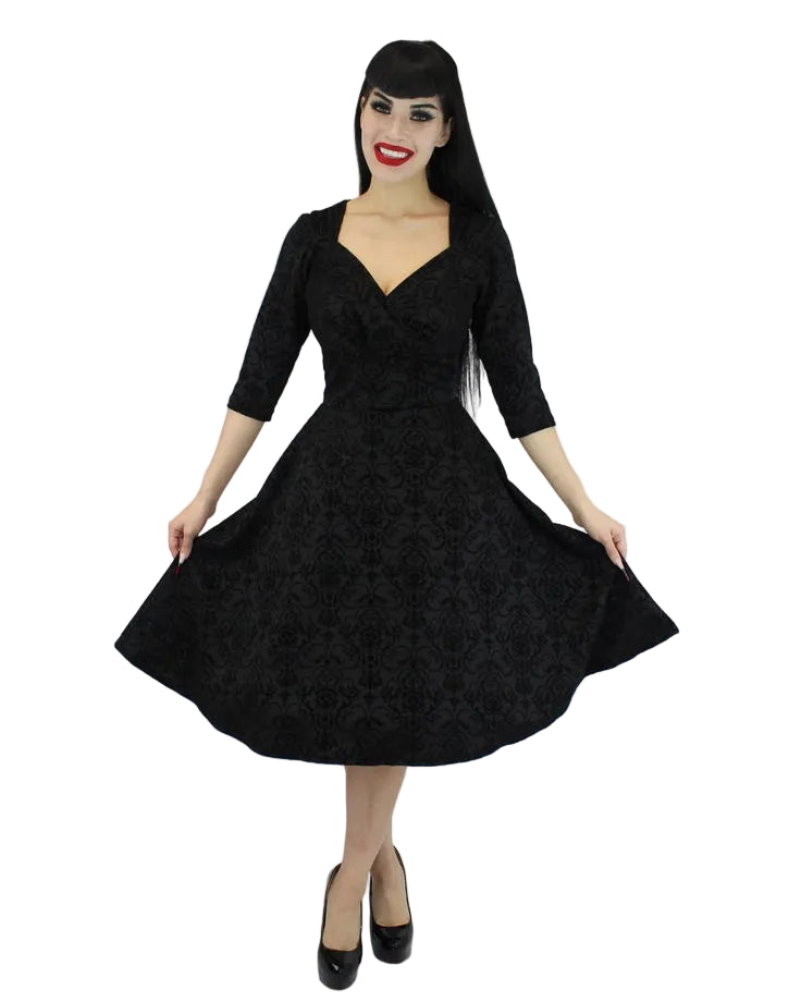 A woman modeling a black 1950s-style swing dress with a textured damask pattern, 3/4 sleeves, and a V-neckline, paired with black heels against a plain white studio background.
