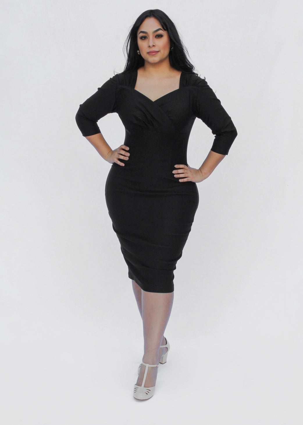 A woman modeling a black knee-length bodycon dress with three-quarter sleeves and a scoop neckline, paired with sheer nude tights and silver high heels, against a light gray studio background.