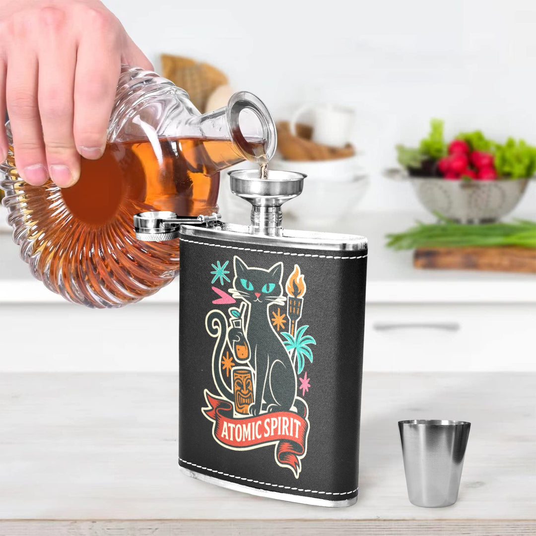 Person pouring liquor into Atomic Spirit black hip flask with spoon and shot glass on a kitchen counter.