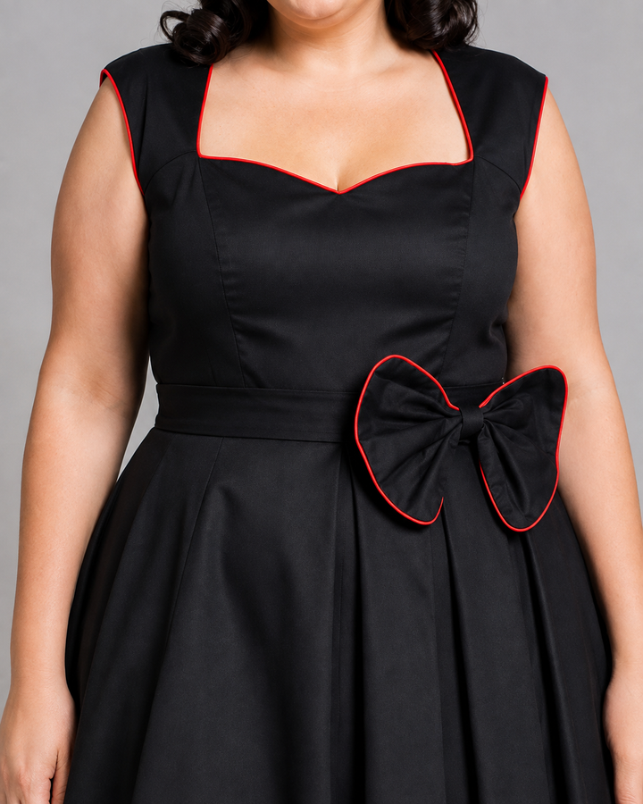 A close-up of a black swing dress with red piping trim, featuring a sweetheart neckline, a defined waist, and a large decorative bow on the side.