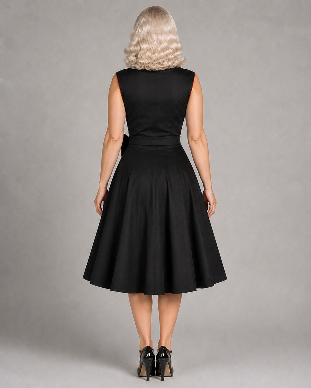 A woman viewed from the back wearing a sleeveless black dress with a fitted waist and full flared skirt, paired with black heels, against a plain gray studio background.