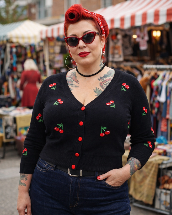Woman wearing a black cardigan with red cherries, sunglasses, and a red bandana at an outdoor market.