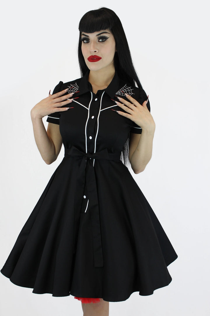 A woman modeling a black retro-style swing dress with white contrast stitching, white buttons, and spiderweb embroidery on the shoulders, set against a plain white background.