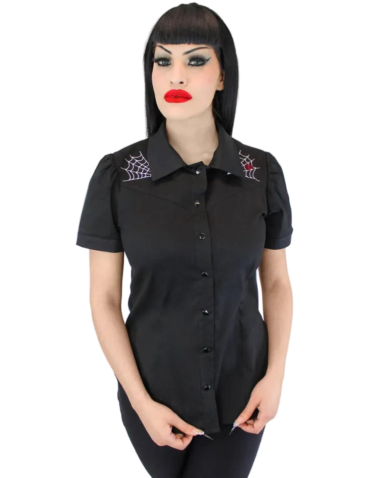 A woman with a black bob haircut and gothic makeup wearing a black short-sleeve button-up shirt with embroidered spiderwebs on the shoulders, set against a plain white background.