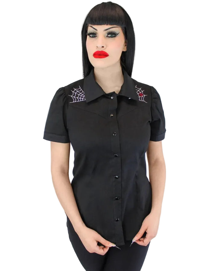 A woman with a black bob haircut and gothic makeup wearing a black short-sleeve button-up shirt with embroidered spiderwebs on the shoulders, set against a plain white background.