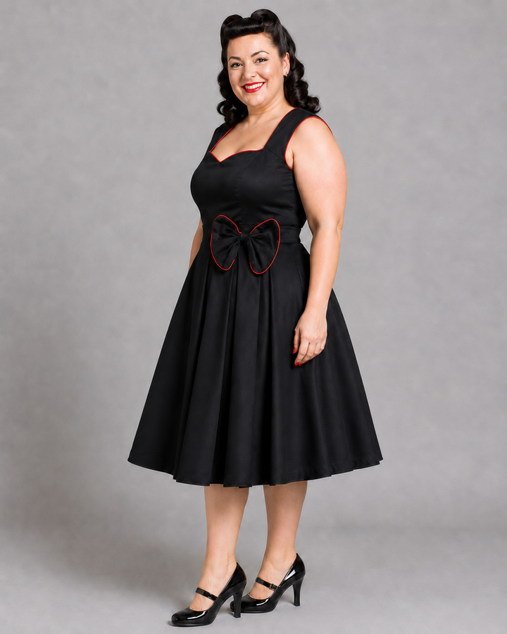A woman modeling a black sleeveless swing dress with a sweetheart neckline, a flared skirt, and a large bow detail at the waist with red trim, paired with black Mary Jane heels against a gray studio background.