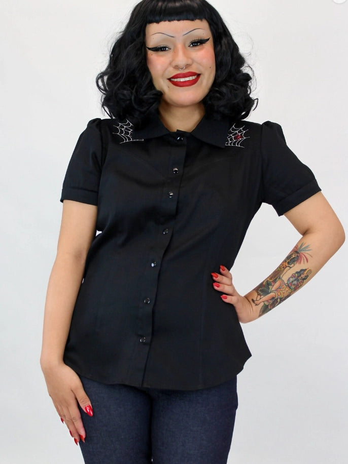 A woman modeling a black short-sleeve button-up blouse with spiderweb embroidery on the shoulders, paired with dark blue jeans, against a plain white background.