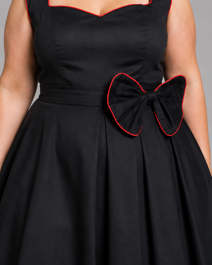 A close-up of a black sleeveless dress with red trim, featuring a structured bodice, a matching waistband, and a large bow detail on the side.