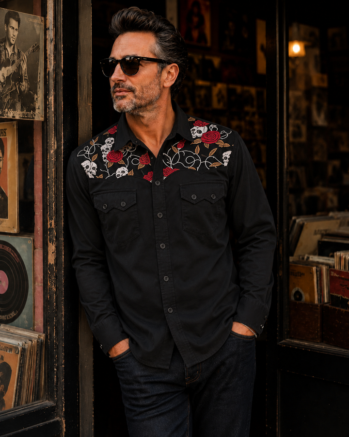 A man wearing a black long-sleeve button-up shirt with embroidered skulls and red roses on the collar, paired with dark jeans and sunglasses, leaning against a rustic door in a dimly lit shop.