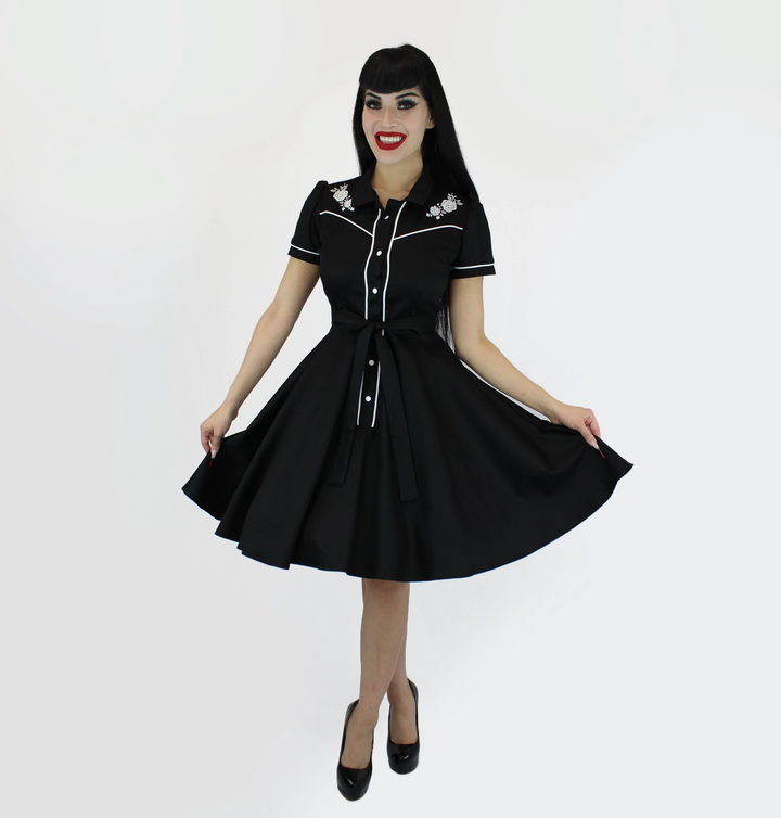 A woman modeling a black 1950s-style swing dress with white piping and floral embroidery on the shoulders, paired with black heels, against a plain white studio background.