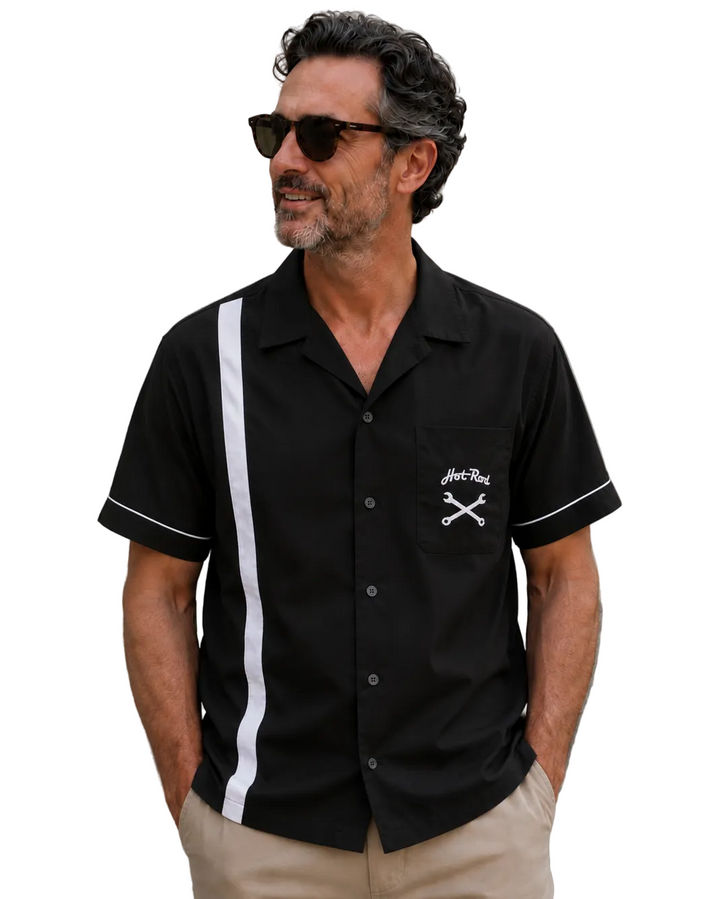 Middle-aged man wearing a black short-sleeve hot rod style bowling shirt with a white vertical stripe, white sleeve piping and chest pocket graphic, standing against a textured concrete wall in beige pants and sunglasses.