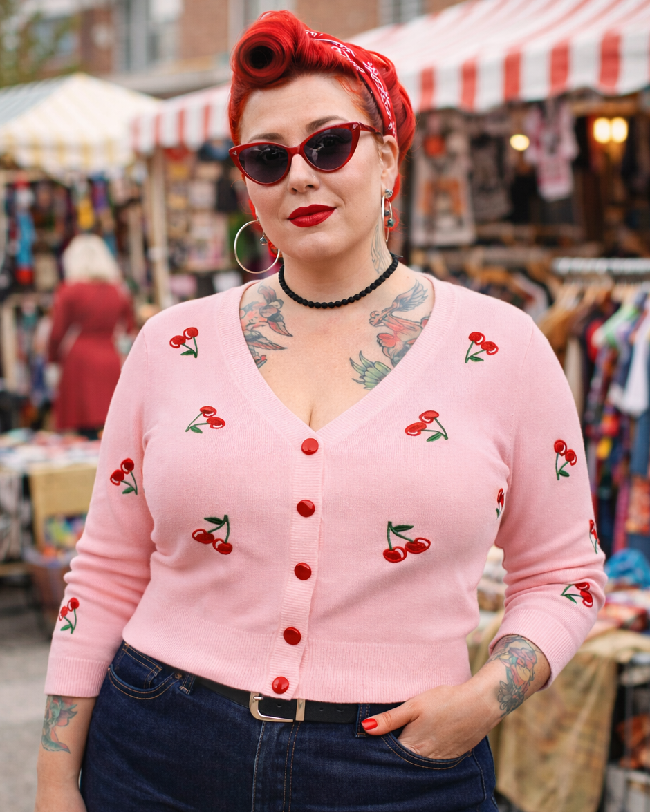 Woman wearing a pink cardigan with cherry designs at an outdoor market.